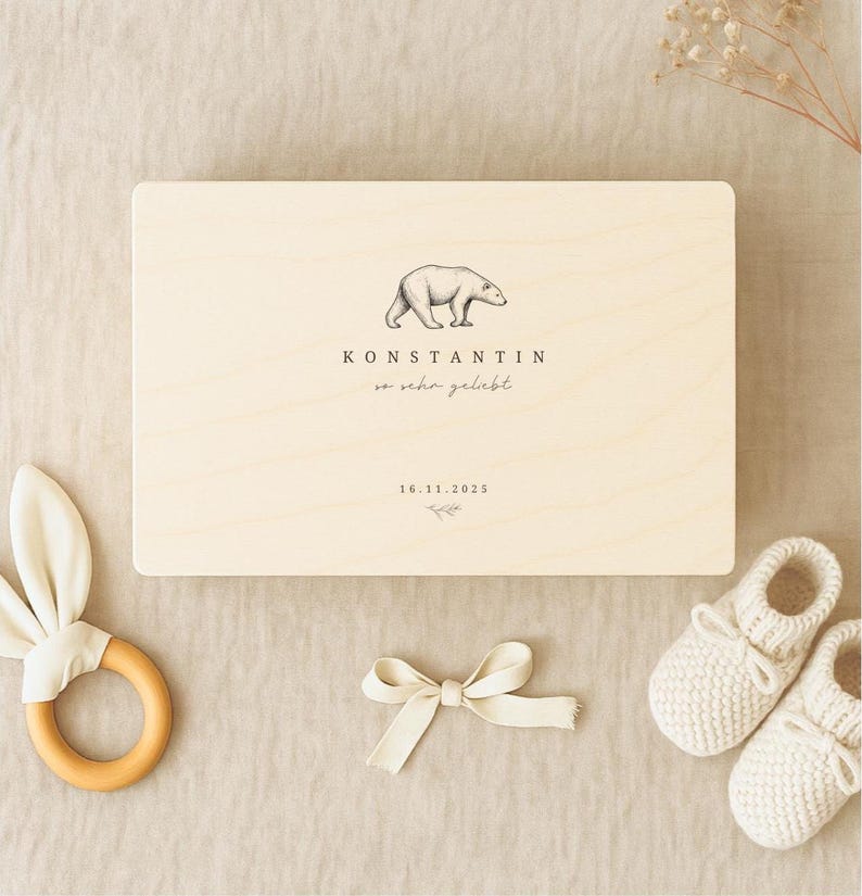 Personalisierte Holz-Erinnerungskiste im Vintage-Bär-Design, flach von oben fotografiert auf beiger Leinentischdecke. Umgeben von Baby-Accessoires wie Holz-Beißring, Stoffschleife und gestrickten Babyschuhen. Name und Datum sind auf dem Deckel aufgedruckt.
