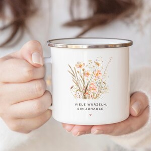 May include: White enamel mug with a silver rim, featuring a watercolor illustration of wildflowers in shades of peach, pink, and beige. The mug has the text "VIELE WURZELN. EIN ZUHAUSE." and a small heart symbol.