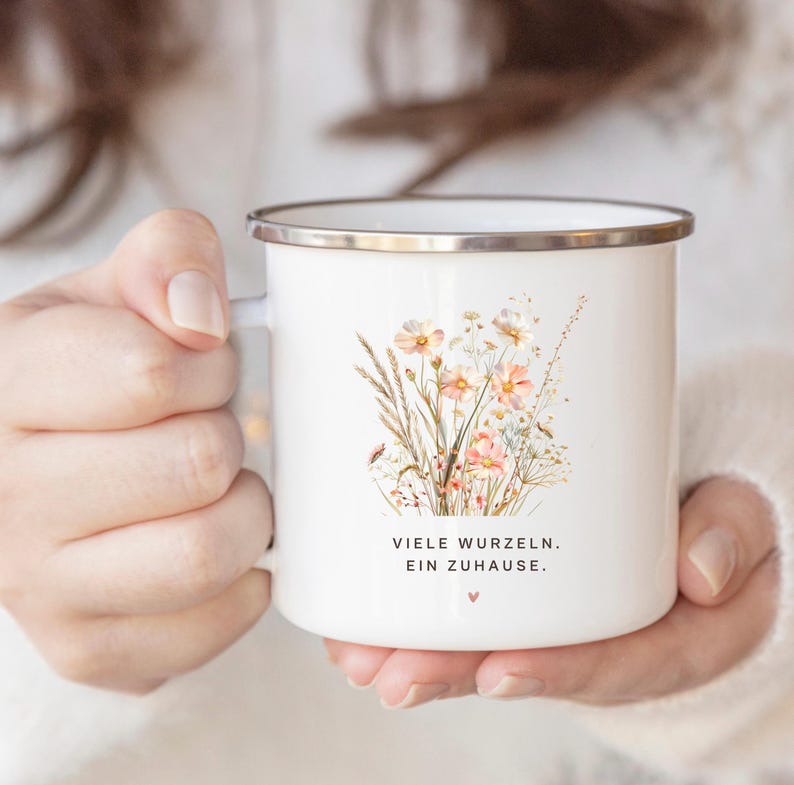 May include: White enamel mug with a silver rim, featuring a watercolor illustration of wildflowers in shades of peach, pink, and beige. The mug has the text "VIELE WURZELN. EIN ZUHAUSE." and a small heart symbol.