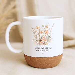 May include: White ceramic mug with a cork base, featuring a floral design in soft pinks and yellows. The mug has a curved handle and the text "VIELE WURZELN. EIN ZUHAUSE." is printed on the front.