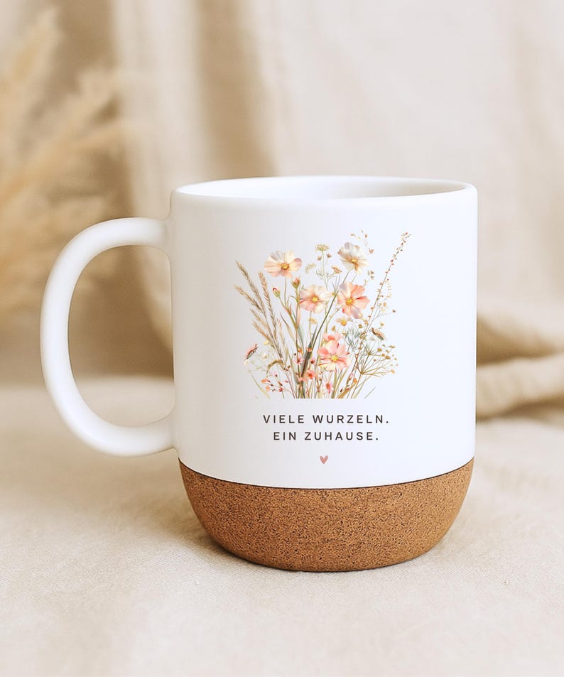 May include: White ceramic mug with a cork base, featuring a floral design in soft pinks and yellows. The mug has a curved handle and the text "VIELE WURZELN. EIN ZUHAUSE." is printed on the front.