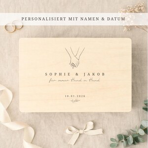 Könnte beinhalten: Eine hellfarbene Holzplakette mit dem Text "SOPHIE & JAKOB" und "für immer Hand in Hand" in zarter Schrift. Die Plakette zeigt eine Strichzeichnung von zwei Händen, die sich halten. Das Datum "10.01.2026" ist ebenfalls enthalten.