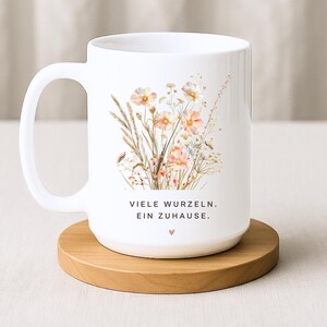 May include: White ceramic mug featuring a floral design with peach and cream-colored flowers and the text "VIELE WURZELN. EIN ZUHAUSE." The mug sits on a round wooden coaster. A heart symbol is below the text.
