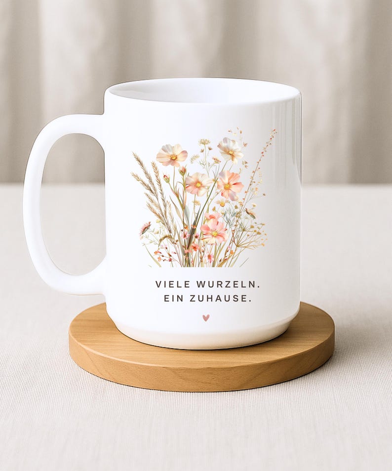 May include: White ceramic mug featuring a floral design with peach and cream-colored flowers and the text "VIELE WURZELN. EIN ZUHAUSE." The mug sits on a round wooden coaster. A heart symbol is below the text.
