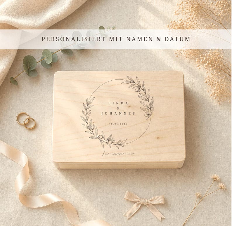 May include: A light-colored wooden box with a circular floral design and the names "Linda & Johannes" and the date "10.01.2026" printed on it. The text "PERSONALISIERT MIT NAMEN & DATUM" is at the top. A gold wedding ring set and a bow are also visible.