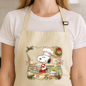 May include: Beige apron featuring a cartoon Snoopy character wearing a chef's hat and apron, baking cookies. The design includes a green kitchen scene with Christmas decorations. The apron has a large front pocket and adjustable neck strap.