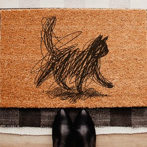 May include: A rectangular doormat with a sketched black cat design. The cat is depicted in a scribbled style, with its tail raised. The mat is brown and sits on a black and white checkered rug. Black shoes are at the bottom.