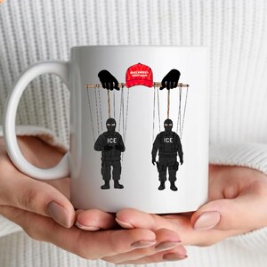 Puede incluir: Taza de cerámica blanca con un diseño de marionetas. El diseño incluye una gorra de béisbol roja con el texto "MAKE AMERICA GREAT AGAIN", manos de marionetas negras y dos figuras con uniformes negros con el texto "ICE".