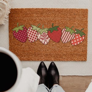 May include: A rectangular doormat with a brown coir surface, decorated with six strawberry designs. Each strawberry has a green stem and a unique pattern, including polka dots, gingham, and stripes. The mat is placed on a light-colored rug.