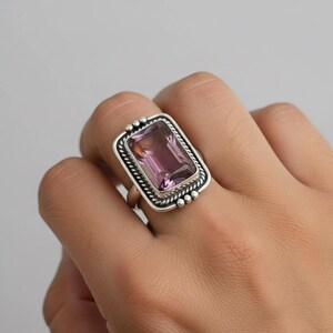 May include: A silver ring with a rectangular amethyst gemstone. The ring has a light purple, rectangular gemstone set within a silver frame. The silver setting has a rope-like border and small decorative beads.