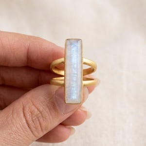 Peut inclure: Une bague dorée ornée d'une pierre précieuse rectangulaire, irisée, bleu clair et blanche. La bague présente un design à double bande avec une monture de pierre verticale, mettant en valeur une esthétique moderne et élégante.