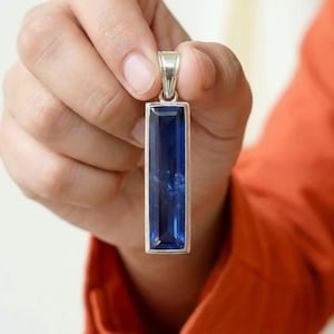 Könnte beinhalten: Ein rechteckiger Anhänger mit einem leuchtend blauen Edelstein, der in einen silberfarbenen Metallrahmen gefasst ist. Der Edelstein hat eine polierte, reflektierende Oberfläche. Der Anhänger wird von einer silbernen Öse gehalten.