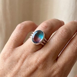 Può includere: Un anello d'argento con una grande pietra preziosa di forma ovale. La pietra preziosa mostra una sfumatura di colori, tra cui blu, viola e rosa. L'anello ha una fascia semplice con tre linee parallele su ogni lato della pietra.
