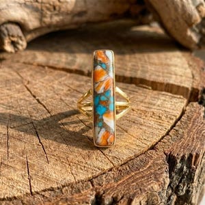 Könnte beinhalten: Ein goldfarbener Ring mit einem rechteckigen, farbenfrohen Stein. Der Stein weist ein Muster aus Orange, Türkis und Weiß auf. Das Band des Rings hat ein geteiltes Design auf jeder Seite, was dem Schmuck eine besondere Note verleiht.