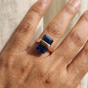 Pode incluir: Um anel de ouro com uma grande pedra preciosa retangular azul escuro. A pedra preciosa é colocada horizontalmente no dedo, com uma simples banda de ouro e uma barra horizontal de ouro que fixa a pedra. O anel é mostrado numa mão.