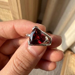 Peut inclure: Une bague en argent ornée d'une grosse pierre précieuse triangulaire rouge foncé. La pierre précieuse facettée capte la lumière, affichant une couleur riche et vibrante. Le corps de la bague a un design ouvert unique. Un bijou.