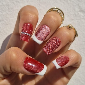 Puede incluir: Primer plano de una mano con esmalte de uñas rojo brillante y puntas blancas. Una uña tiene una banda de pedrería plateada. Se ven dos anillos de oro en los dedos. Las uñas son de forma cuadrada.