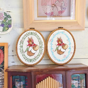 May include: Two oval-shaped embroidered hoops featuring a rabbit in a red cape and holding a basket and a green leaf. One hoop has a green floral border, the other a blue floral border. Both are set in wooden frames.