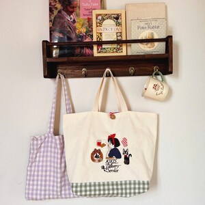 Pode incluir: Uma sacola creme com um design bordado com uma menina, um gato e o texto "Kiki's Delivery Service". Uma sacola xadrez roxa pendurada &agrave; esquerda. Uma prateleira de madeira cont&eacute;m livros e uma caneca.