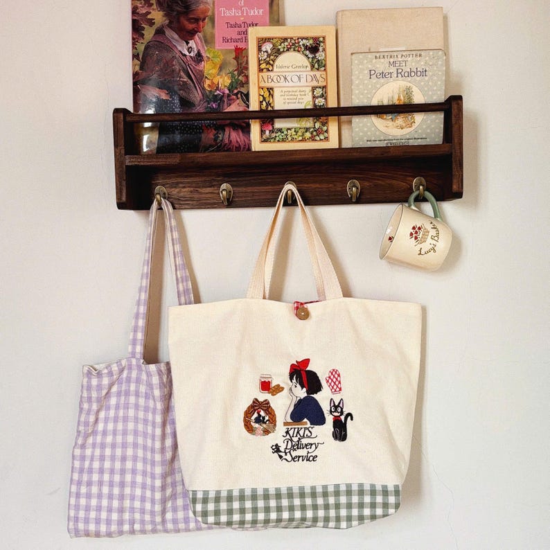Pode incluir: Uma sacola creme com um design bordado com uma menina, um gato e o texto "Kiki's Delivery Service". Uma sacola xadrez roxa pendurada &agrave; esquerda. Uma prateleira de madeira cont&eacute;m livros e uma caneca.
