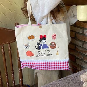 K&ouml;nnte beinhalten: Beige Tragetasche mit gestickten Illustrationen und dem Text "KIKI'S DELIVERY SERVICE". Die Tasche hat einen rot-wei&szlig; karierten Boden, einen Knopfverschluss und ein gestreiftes Innenfutter. Das Design umfasst eine schwarze Katze, ein M&auml;dchen mit roter Schleife und andere thematische Grafiken.