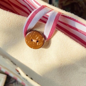 K&ouml;nnte beinhalten: Nahaufnahme einer beigefarbenen Canvas-Tragetasche mit einem rot-wei&szlig; gestreiften Bandgriff und Innenfutter. Ein runder, brauner Knopf aus Kokosnussschale sichert die &Ouml;ffnung der Tasche. Die Tasche hat eine schlichte, nat&uuml;rliche &Auml;sthetik.