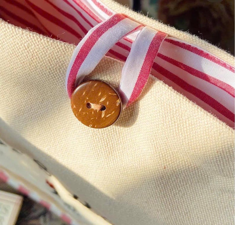 K&ouml;nnte beinhalten: Nahaufnahme einer beigefarbenen Canvas-Tragetasche mit einem rot-wei&szlig; gestreiften Bandgriff und Innenfutter. Ein runder, brauner Knopf aus Kokosnussschale sichert die &Ouml;ffnung der Tasche. Die Tasche hat eine schlichte, nat&uuml;rliche &Auml;sthetik.