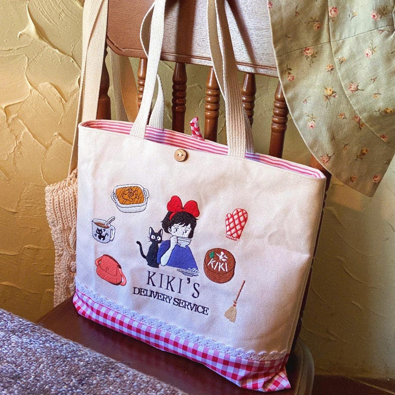 K&ouml;nnte beinhalten: Cremefarbene Tragetasche mit gestickten Illustrationen aus Kiki's Delivery Service. Die Tasche hat einen rot-wei&szlig; karierten Besatz, einen Knopfverschluss und cremefarbene Griffe. Das Design zeigt Kiki, Katze Jiji und Essen.