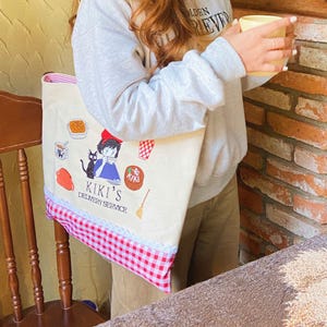 K&ouml;nnte beinhalten: Eine beigefarbene Tragetasche mit einer Cartoon-Illustration und dem Text "Kiki's Delivery Service". Die Tasche hat unten ein rot-wei&szlig;es Karomuster mit einem rosa Besatz. Die Tasche wird von einer Person gehalten.