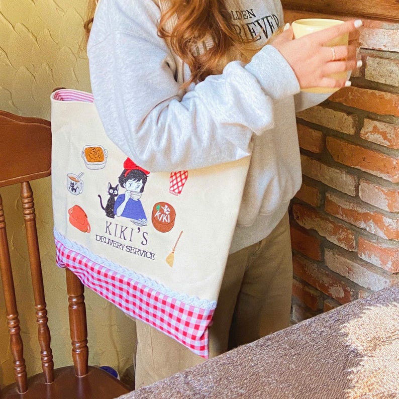 K&ouml;nnte beinhalten: Eine beigefarbene Tragetasche mit einer Cartoon-Illustration und dem Text "Kiki's Delivery Service". Die Tasche hat unten ein rot-wei&szlig;es Karomuster mit einem rosa Besatz. Die Tasche wird von einer Person gehalten.