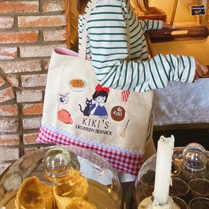 K&ouml;nnte beinhalten: Eine beigefarbene Tragetasche mit rot-wei&szlig;em Vichy-Besatz und wei&szlig;em Spitzenrand. Die Tasche zeigt gestickte Illustrationen und den Text "KIKI'S DELIVERY SERVICE". Eine Person tr&auml;gt ein gr&uuml;n-wei&szlig; gestreiftes Langarmshirt.