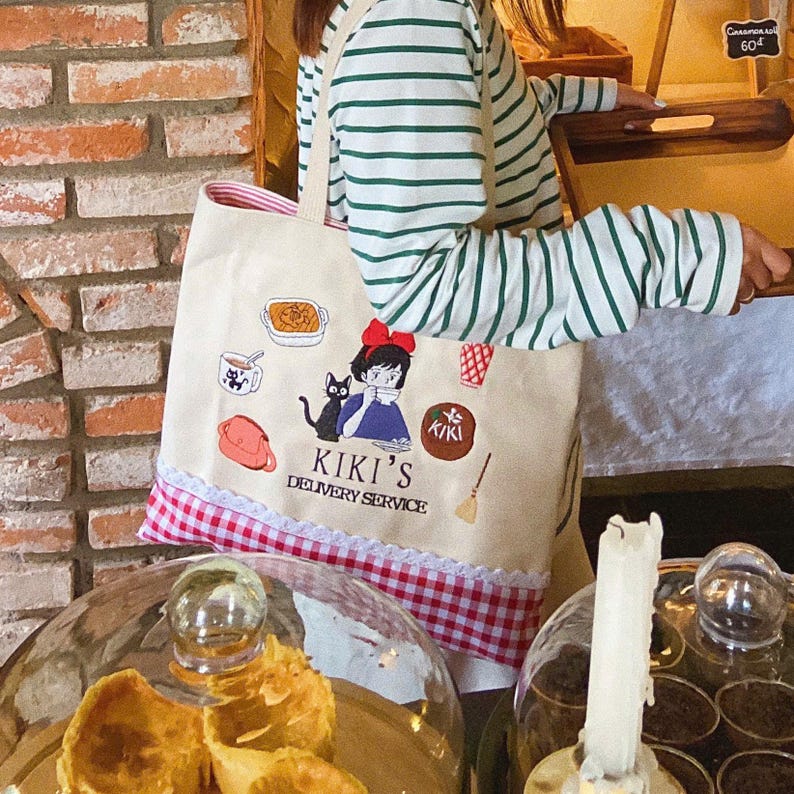 K&ouml;nnte beinhalten: Eine beigefarbene Tragetasche mit rot-wei&szlig;em Vichy-Besatz und wei&szlig;em Spitzenrand. Die Tasche zeigt gestickte Illustrationen und den Text "KIKI'S DELIVERY SERVICE". Eine Person tr&auml;gt ein gr&uuml;n-wei&szlig; gestreiftes Langarmshirt.