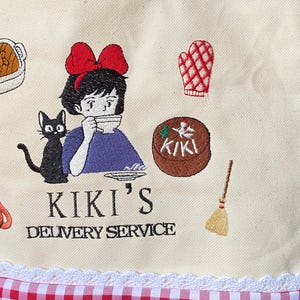 K&ouml;nnte beinhalten: Cremefarbener Stoff mit einem gestickten Design. Es zeigt ein M&auml;dchen mit roter Schleife, eine schwarze Katze und den Text "KIKI'S DELIVERY SERVICE". Weitere gestickte Elemente sind ein Kuchen, ein Ofenhandschuh und ein Besen.