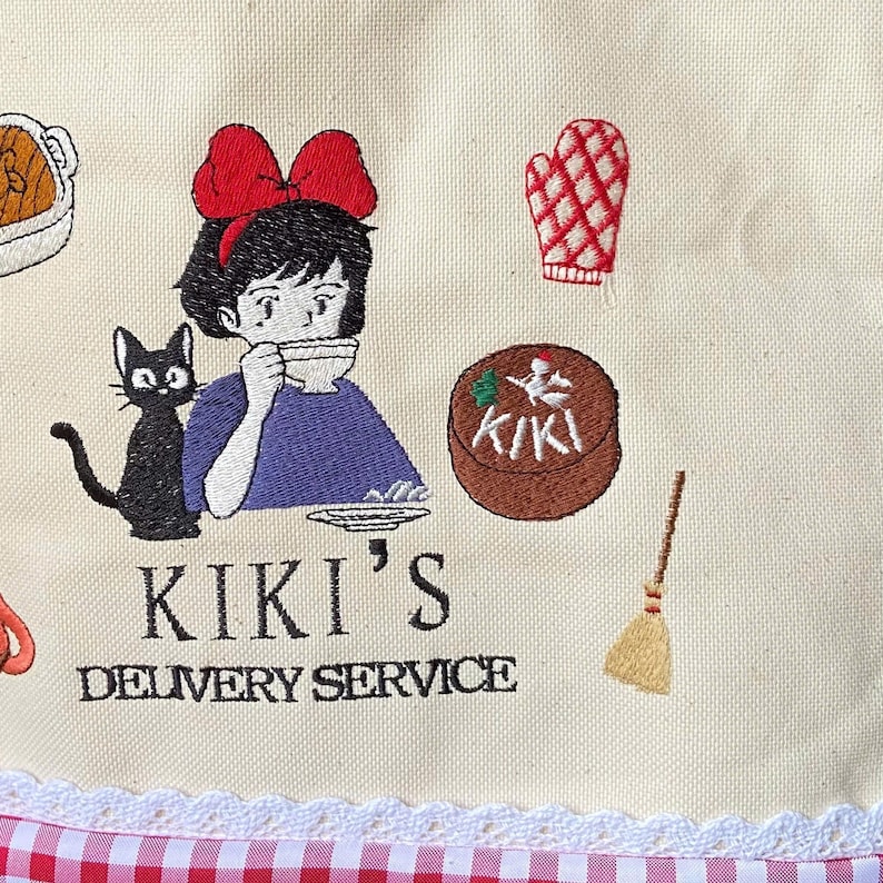 K&ouml;nnte beinhalten: Cremefarbener Stoff mit einem gestickten Design. Es zeigt ein M&auml;dchen mit roter Schleife, eine schwarze Katze und den Text "KIKI'S DELIVERY SERVICE". Weitere gestickte Elemente sind ein Kuchen, ein Ofenhandschuh und ein Besen.