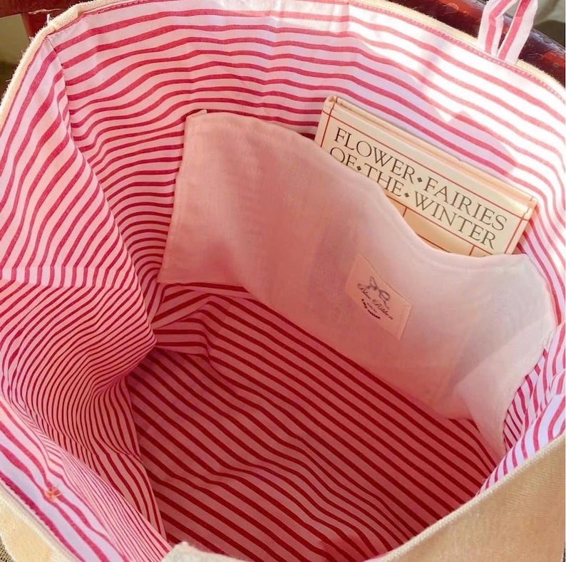 K&ouml;nnte beinhalten: Beige Tragetasche mit rot-wei&szlig; gestreiftem Futter. Eine hellrosa Tasche ist im Inneren der Tasche sichtbar, zusammen mit einem Buch mit dem Titel "Flower Fairies of the Winter". Die Tasche hat ein schlichtes Design und ist f&uuml;r den t&auml;glichen Gebrauch geeignet.
