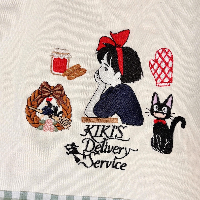 Pode incluir: Design bordado em tecido creme com uma menina com la&ccedil;o vermelho, um gato preto e o texto "Kiki's Delivery Service". Outros elementos incluem um pote de geleia, p&atilde;o, uma coroa de flores e uma luva de forno vermelha.