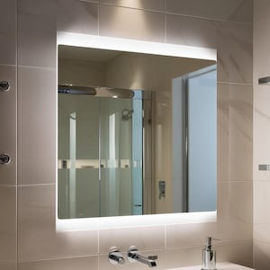 Può includere: Uno specchio da bagno quadrato e illuminato con un bordo a LED bianco brillante. Lo specchio è montato su una parete piastrellata sopra un lavandino bianco con accessori cromati. Il riflesso mostra una doccia e altri elementi del bagno.