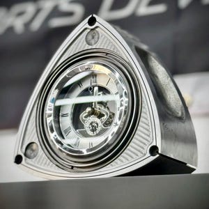 Puede incluir: Un rotor de motor rotativo de color plateado reutilizado como reloj de escritorio. La carcasa metálica triangular presenta una esfera circular con números romanos negros, manecillas plateadas y un mecanismo interno visible tras un cristal.