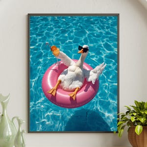 Puede incluir: Impresión enmarcada de un ganso blanco con gafas de sol flotando en un flotador rosa en una piscina. El ganso sostiene un cóctel y está rodeado de agua azul. La imagen está enmarcada en negro.
