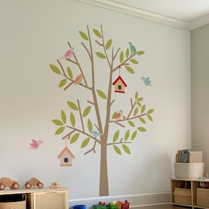 May include: A wall decal featuring a tree with green leaves, brown branches, and colorful birds. Two birdhouses are also included. The decal is applied to a light-colored wall in a child's room. Wooden toy cars and storage baskets are in the foreground.