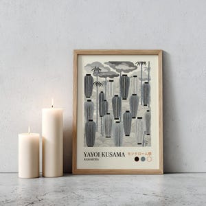 May include: A framed print featuring a black and white illustration of Japanese lanterns, bamboo, and clouds. The print includes the text "YAYOI KUSAMA KAMAKURA" in black and Japanese characters. Two white pillar candles are lit in front of the print.