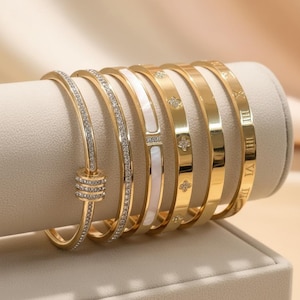 Può includere: Una collezione di braccialetti color oro esposti su un supporto color crema. I braccialetti presentano vari design, tra cui dettagli di diamanti, numeri romani e motivi floreali. I braccialetti sono realizzati in un materiale lucido e riflettente.