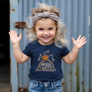 May include: A young child wearing a navy blue t-shirt with a graphic of a creature and the text "YOU SLEEP. I WATCH." The child is also wearing blue jeans and a tan headband. The background is a blue corrugated metal wall.