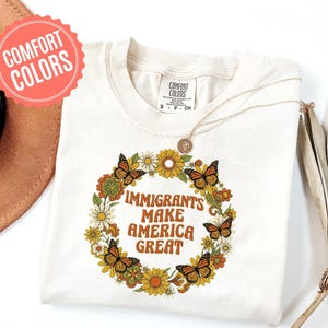 以下が含まれることがあります： クリーム色のTシャツ。花と蝶のリースデザイン。リースはオレンジ色の「IMMIGRANTS MAKE AMERICA GREAT」という文字を囲んでいます。Tシャツは丸いネックラインと半袖です。金のネックレスがTシャツの上にかけられています。