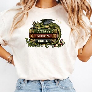 Può includere: Una t-shirt color crema con la grafica di un drago verde che riposa su una pila di tre libri. I dorsi dei libri riportano le scritte FANTASY, DYSTOPIAN e THRILLER in lettere dorate. Il disegno include foglie di edera e piccoli fiori.