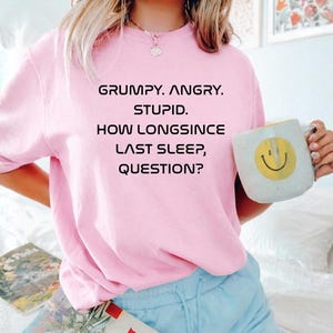 May include: A light pink sweatshirt with the text "GRUMPY. ANGRY. STUPID. HOW LONGSINCE LAST SLEEP, QUESTION?" in black. A white mug with a yellow smiley face is held in the hand. The sweatshirt is paired with light blue shorts.