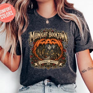 Può includere: Una t-shirt grigio scuro mélange con una stampa grafica di una grande zucca arancione che incornicia una scena di strada buia con edifici, una luna crescente e un gatto nero. Il testo recita "MIDNIGHT BOOKTOWN LITERARY SOCIETY EST. 2026".