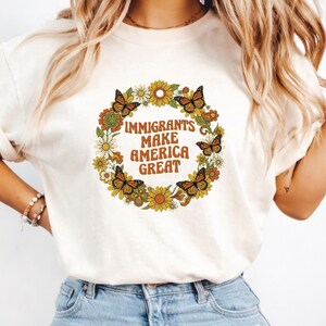 以下が含まれることがあります： クリーム色のTシャツ。蝶と「IMMIGRANTS MAKE AMERICA GREAT」の文字が茶色とオレンジ色で描かれた花のリースデザイン。デザインには黄色と白の花が含まれています。