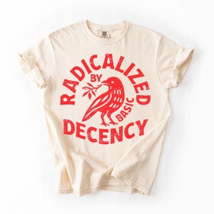 Peut inclure: T-shirt crème avec un motif rouge. Le motif représente un oiseau tenant une branche et les mots "RADICALIZED BY BASIC DECENCY" disposés en cercle. Le t-shirt a des manches retroussées.