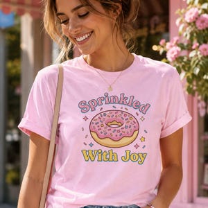 Könnte beinhalten: Ein rosa T-Shirt mit hochgekrempelten Ärmeln, das eine Grafik eines Donuts mit rosa Streuseln und dem Text "Sprinkled With Joy" zeigt. Das Shirt wird mit einer blauen Jeans kombiniert.